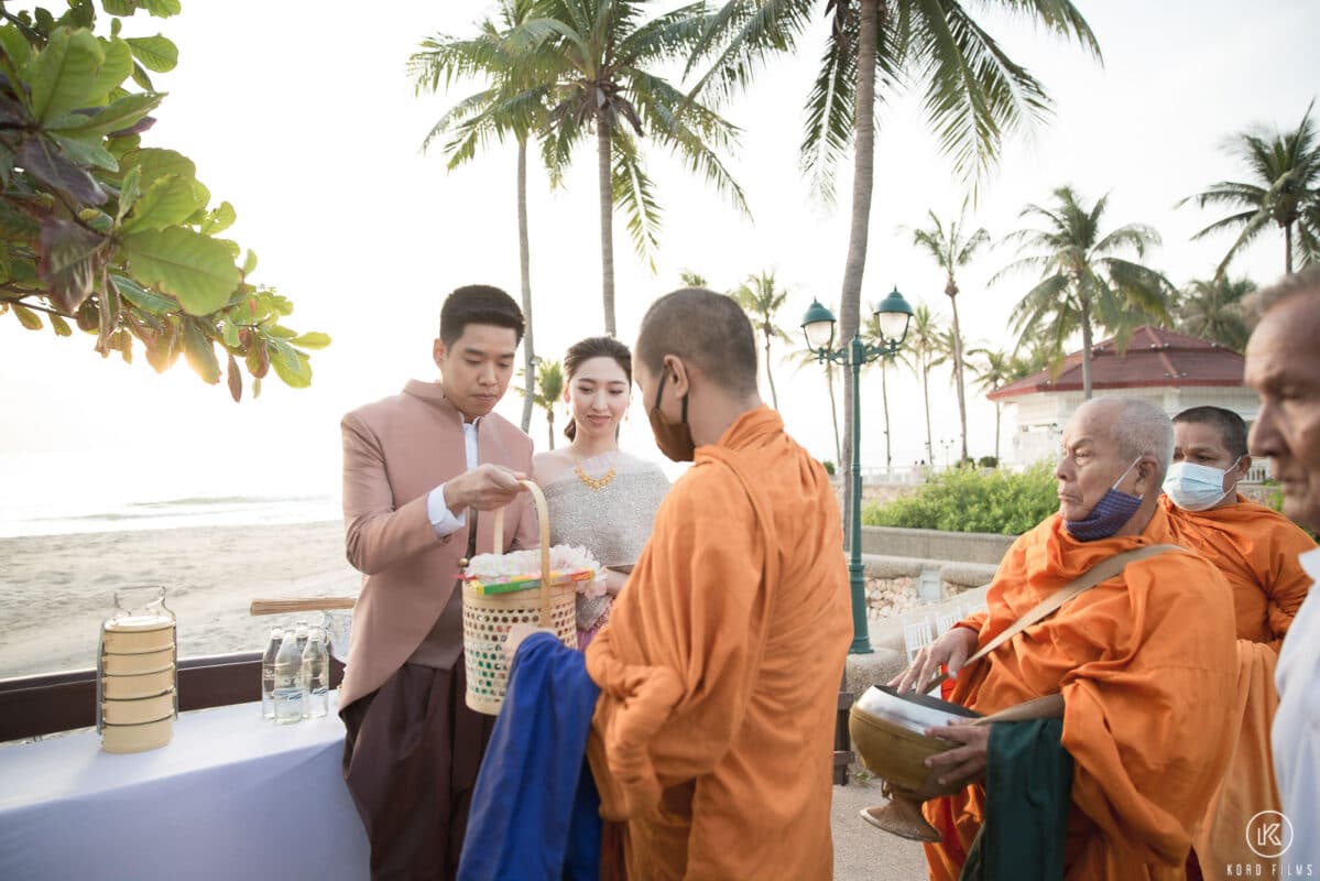 Bride and groom in Thai silk outfits offer food to Buddhist monks at sunrise on the beach at Dusit Thani Hua Hin
