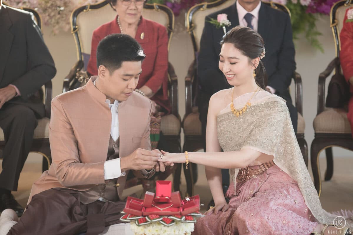 The groom slips the engagement ring onto the bride's finger as both families watch at Dusit Thani Hua Hin