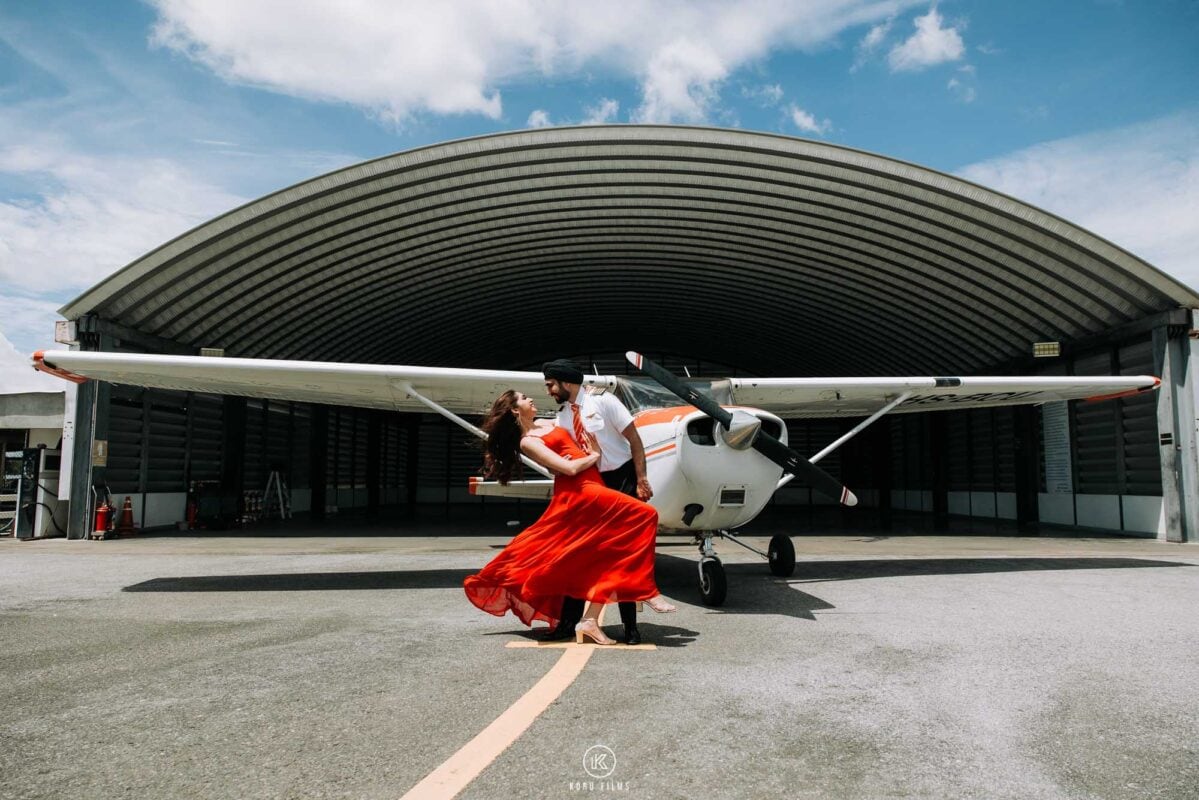 Ritu in a flowing red gown and Naveen in front of a small propeller plane at an airport hangar