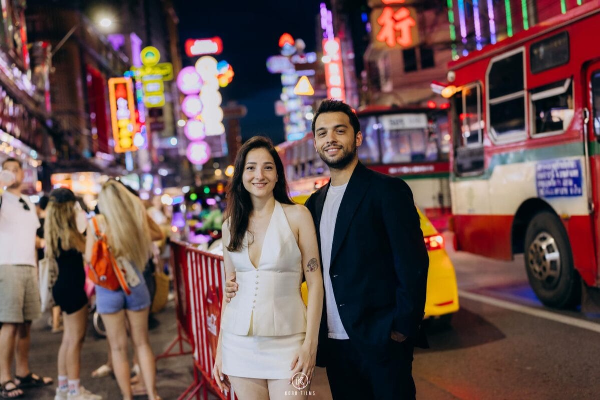 Couple stand on Yaowarat Road at night under neon signs, with a red bus behind them, Chinatown, Bangkok