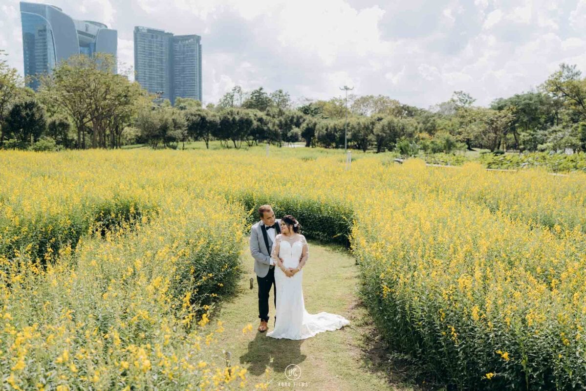 Couple in a white gown and grey suit in a field of yellow flowers with Bangkok towers behind, Rod Fai Park
