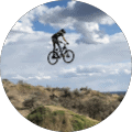 A person on a mountain bike is airborne above a dirt trail with hills and a cloudy sky in the background.