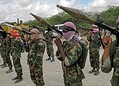 Line of masked soldiers in camouflage uniforms aiming shoulder-fired weapons with large rocket launchers in a dusty outdoor area.