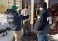 Two officers in HSI jackets speak with a man at a store entrance on a snowy sidewalk, near a glass door with an 'OPEN' sign visible.