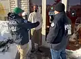 Two officers in HSI jackets speak with a man at a store entrance on a snowy sidewalk, near a glass door with an 'OPEN' sign visible.