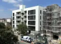 Contemporary white apartment building with large black framed windows and open balconies, modern minimalist style, under construction.