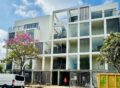 Modern white multi-story apartment building with open-air corridors and balconies, featuring vertical slat accents and a geometric facade.
