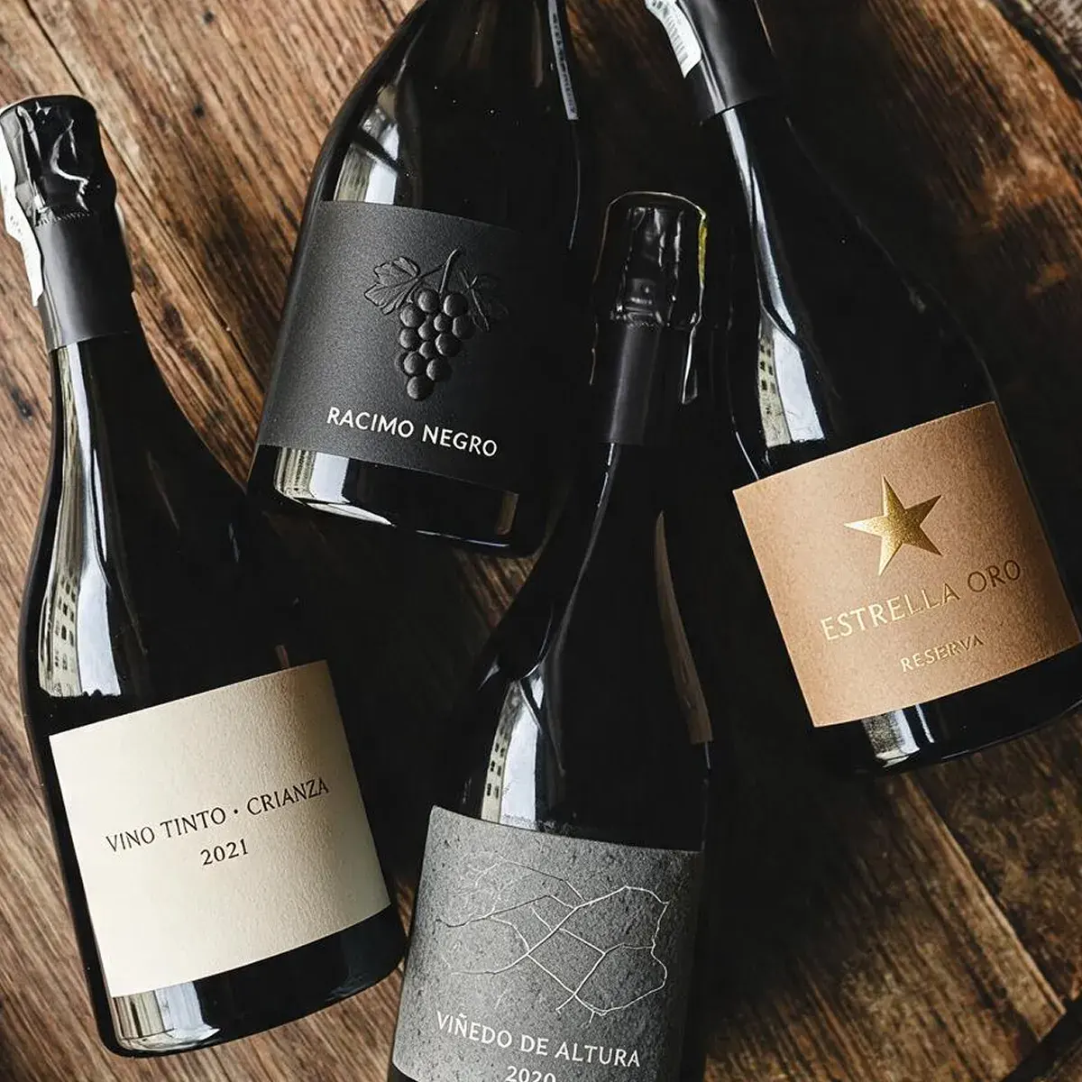 Cuatro botellas de vino mostrando distintos diseños y acabados de etiquetas personalizadas, incluyendo estampados en relieve, texturas de piedra y detalles dorados, sobre una superficie de madera.