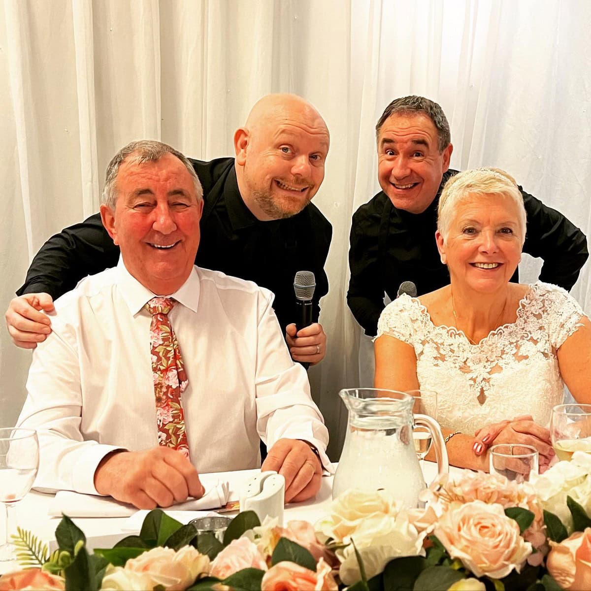 The Sing Along Waiters surprising mature newlyweds at elegant UK wedding reception, award-winning entertainment duo with 30 years experience, featuring peach rose table decorations and intimate venue setting, premium North West wedding entertainment specialists