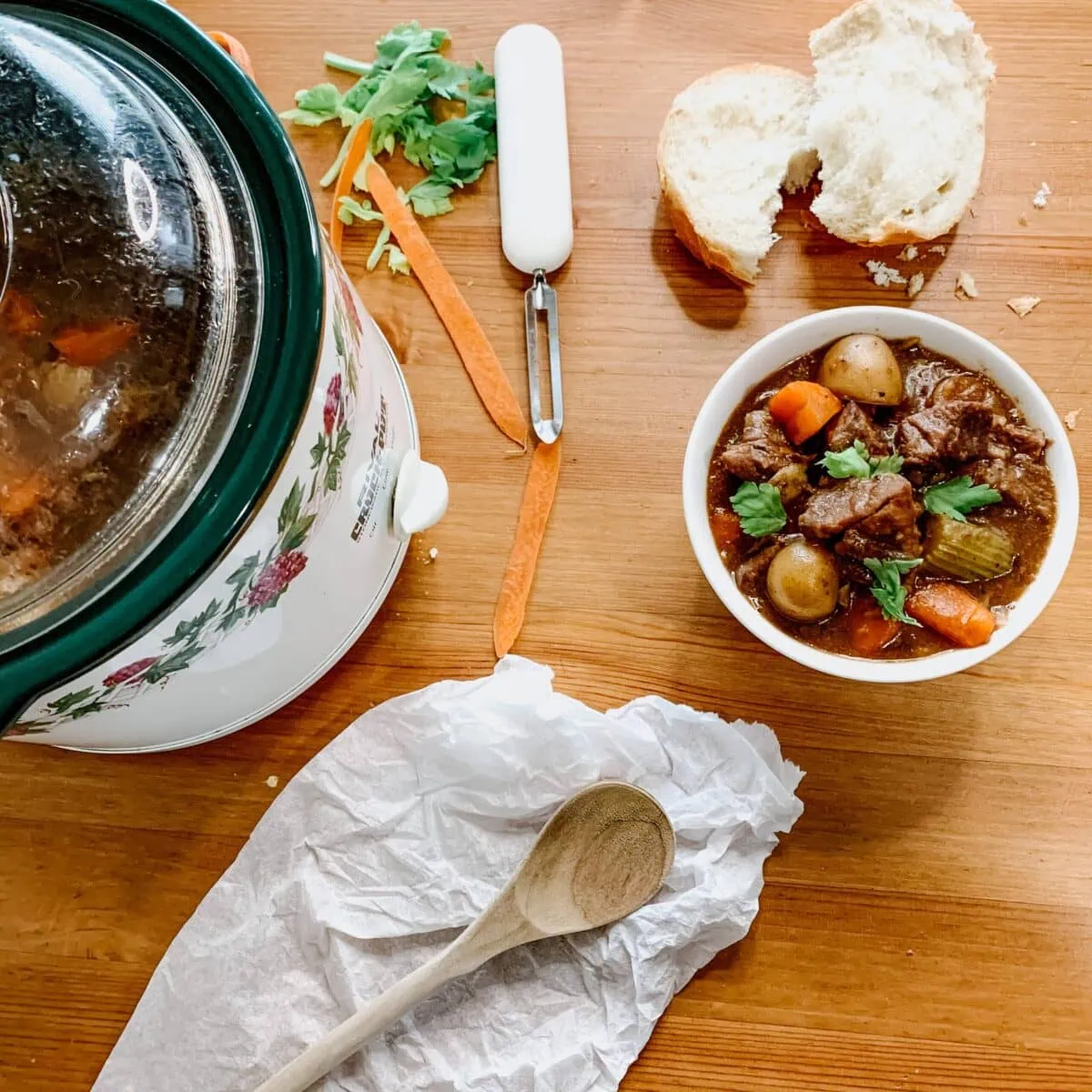 beef stew in a bowl, bread, carrots and parsley on the side, a crock pot on the side