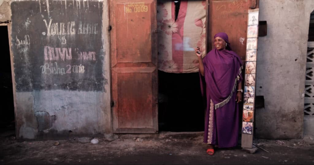 Scene from Apostles of Cinema (2023) showing a woman in a purple dress smiling outside a weathered building, embodying joy and community storytelling in East Africa.