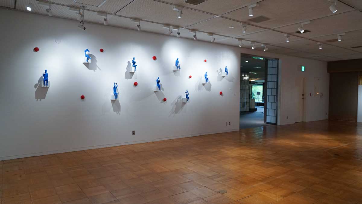Kazumasa Mizokami Exhibition scene 5　Nine small blue figures are lined up on a white wall, with small red flowers between them.