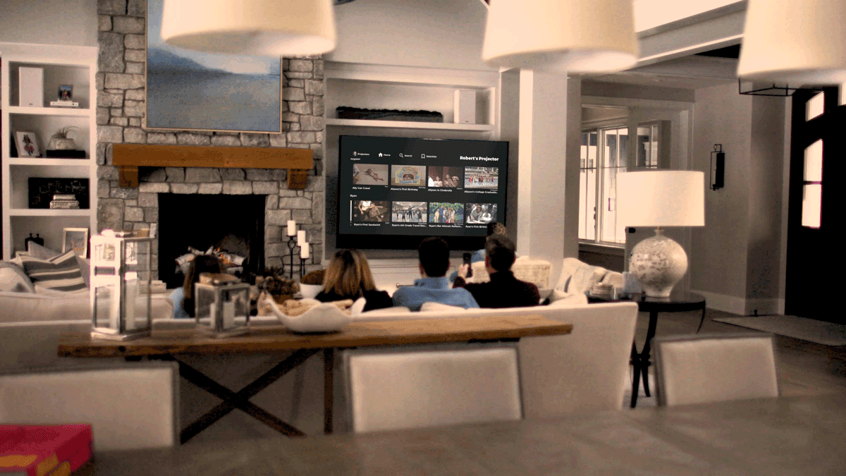 A family of four sits on a sofa in a cozy living room watching a movie on a large TV mounted above a stone fireplace. The room is softly lit with modern decor, including a large table, lamps, and shelves filled with various items.
