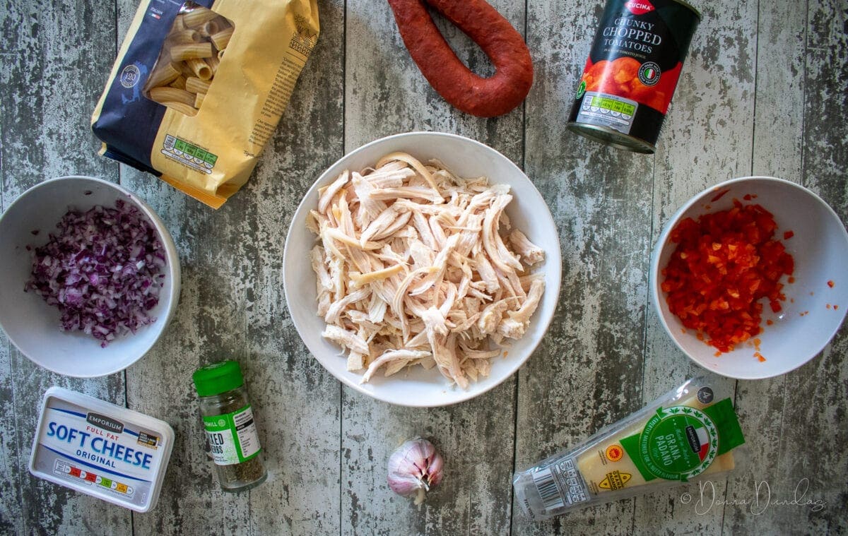 ingredients for leftover roast chicken pasta laid on table. Shredded chicken, diced pepper, diced onion, pasta in pack, chorizo, tin of chopped tomatoes, parmesan, garlic build, mixed herbs and soft cheese.