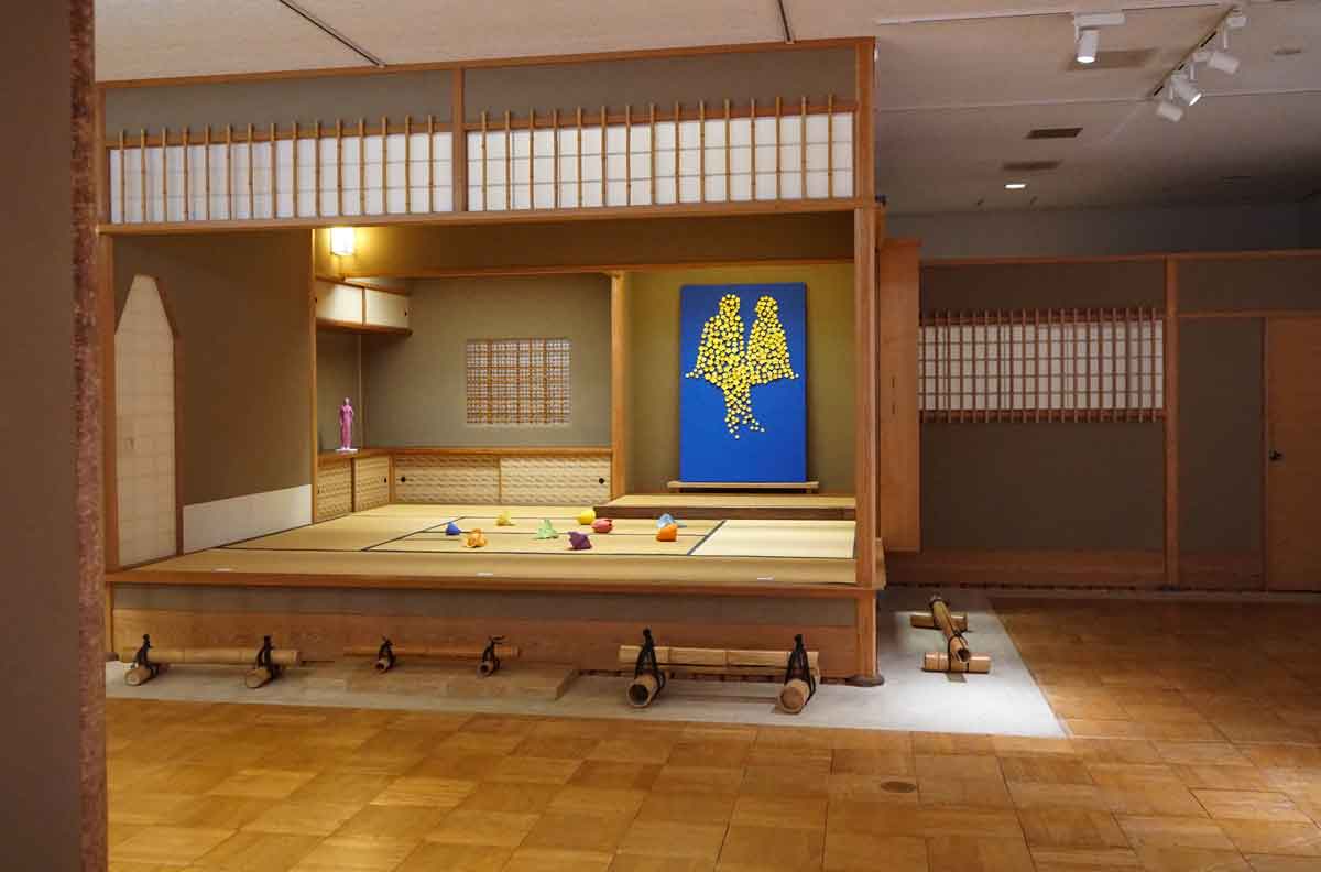 Kazumasa Mizokami Exhibition scene 7　There is a tea room set up inside the museum, and at the back, a yellow figure of lovers is floating above a blue canvas.