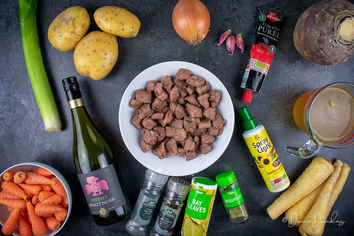 ingredients for lamb stew - cooked lamb diced in white bowl, onion, 3 cloves of garlic, tube of tomato puree, whole swede, stock in jug, bottle of cooking spray, 4 parsnips, small tub of rosemary, tub of bay leaves, salt and pepper in grinders, bottle of red wine, chantenay carrots in white bowl, large leek, 3 baking potatoes.