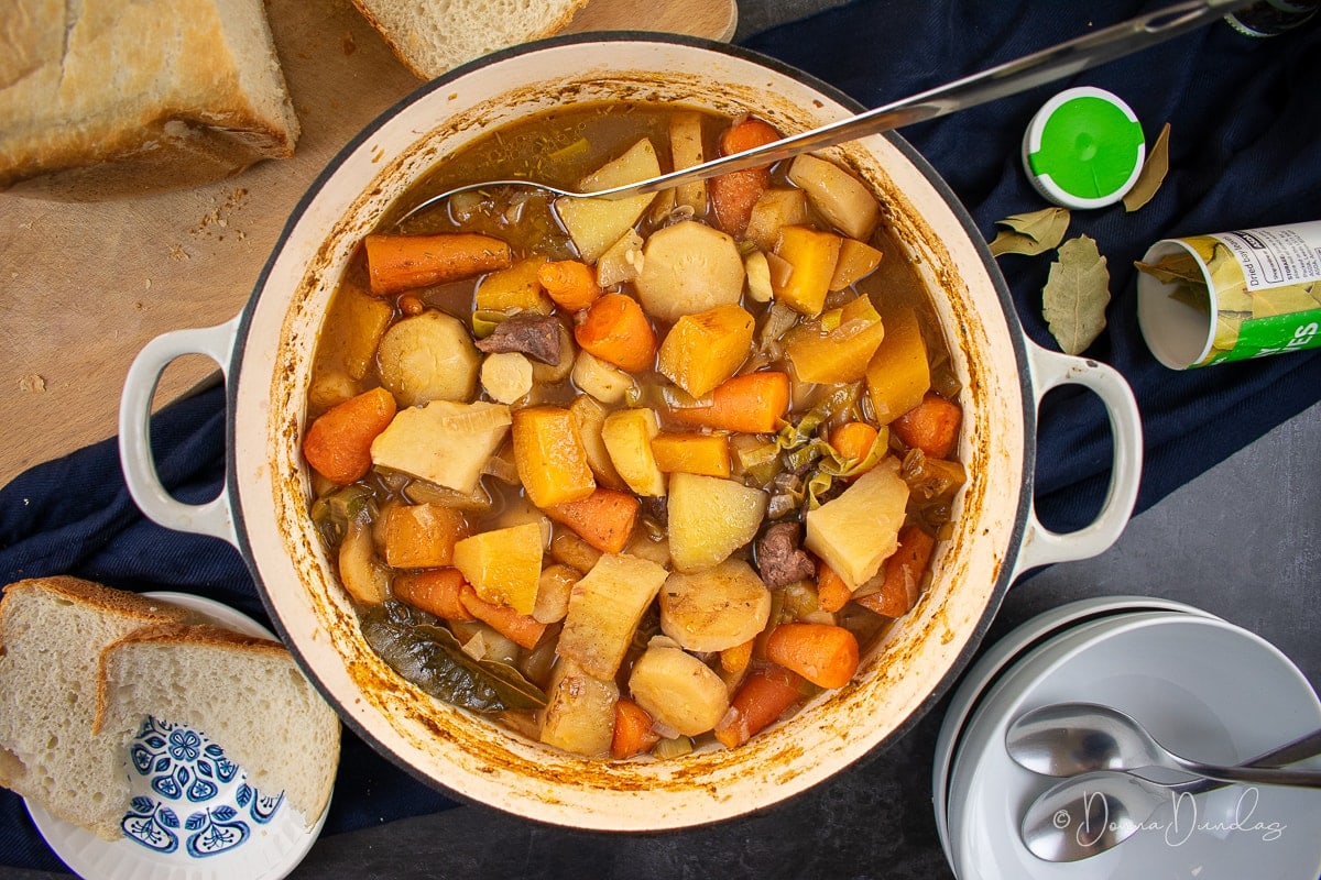 stew in cast iron dish. Loaf of bread, bay leaves, salt and pepper and empty bowls to the sides.