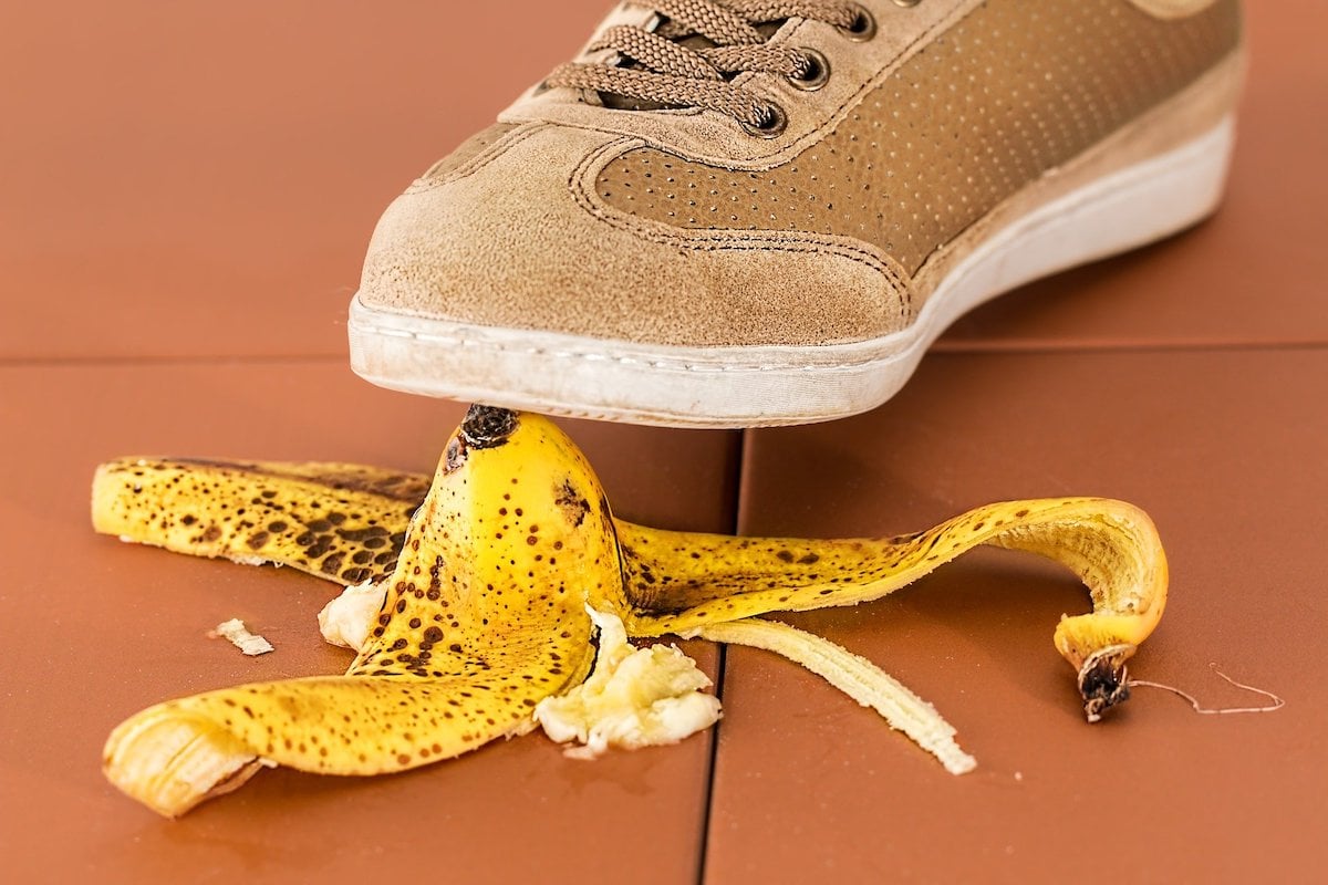 Schuh tritt auf eine braun gesprenkelte Bananenschale auf einem braunen Fliesenboden, symbolisiert eine Gefahrensituation.