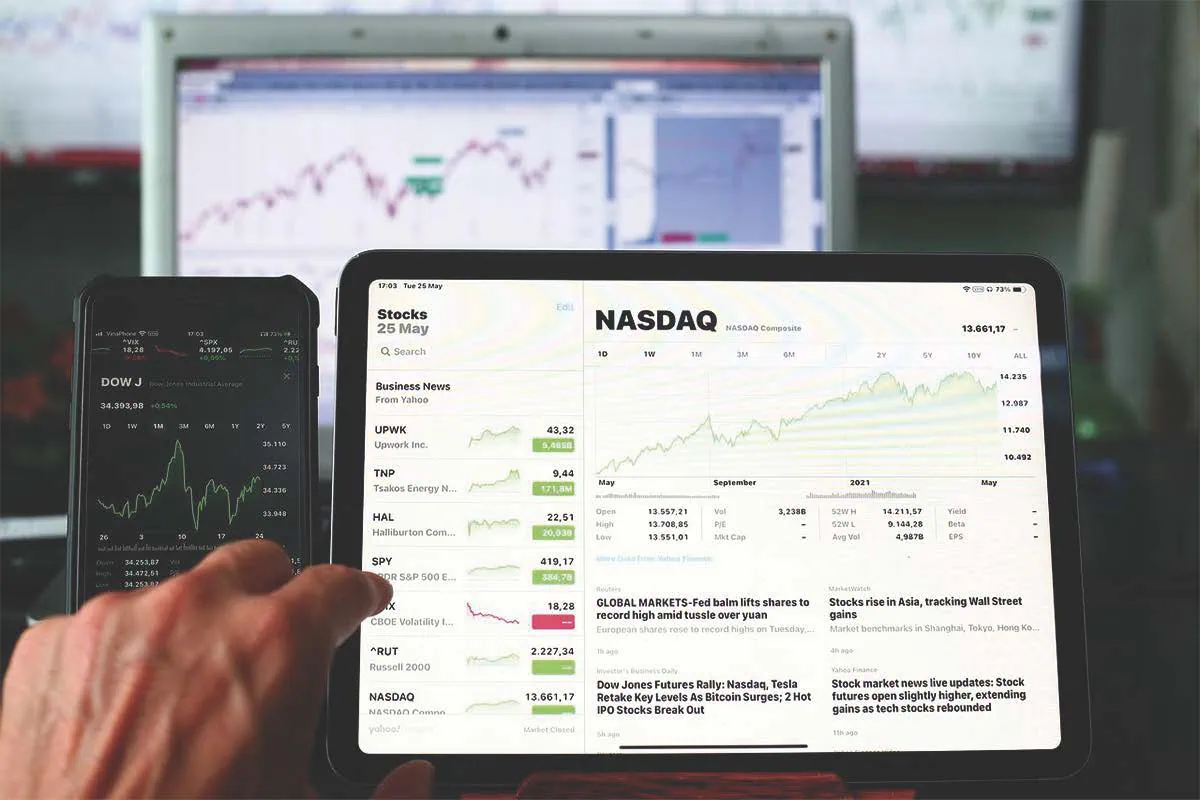 Hand zeigt auf Tablet mit NASDAQ-Chart; daneben Smartphone mit Dow Jones und Kursdaten.