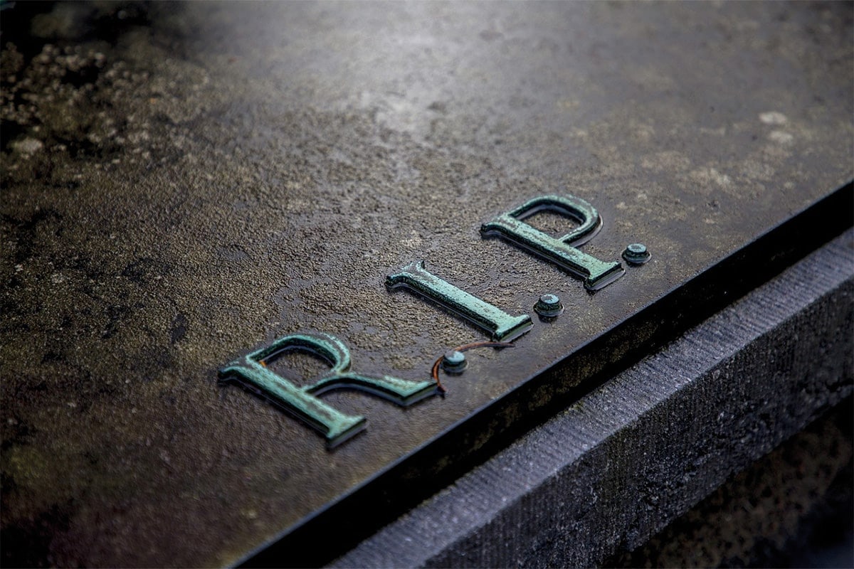 Nahaufnahme eines Grabsteins mit erhabener, grüner R.I.P.-Inschrift auf dunklem Stein.
