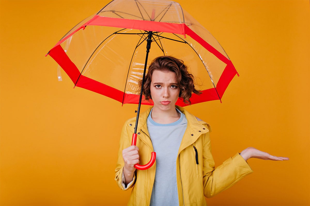 Frau mit gelber Regenjacke und rotem Regenschirm blickt skeptisch, vor orangem Hintergrund.