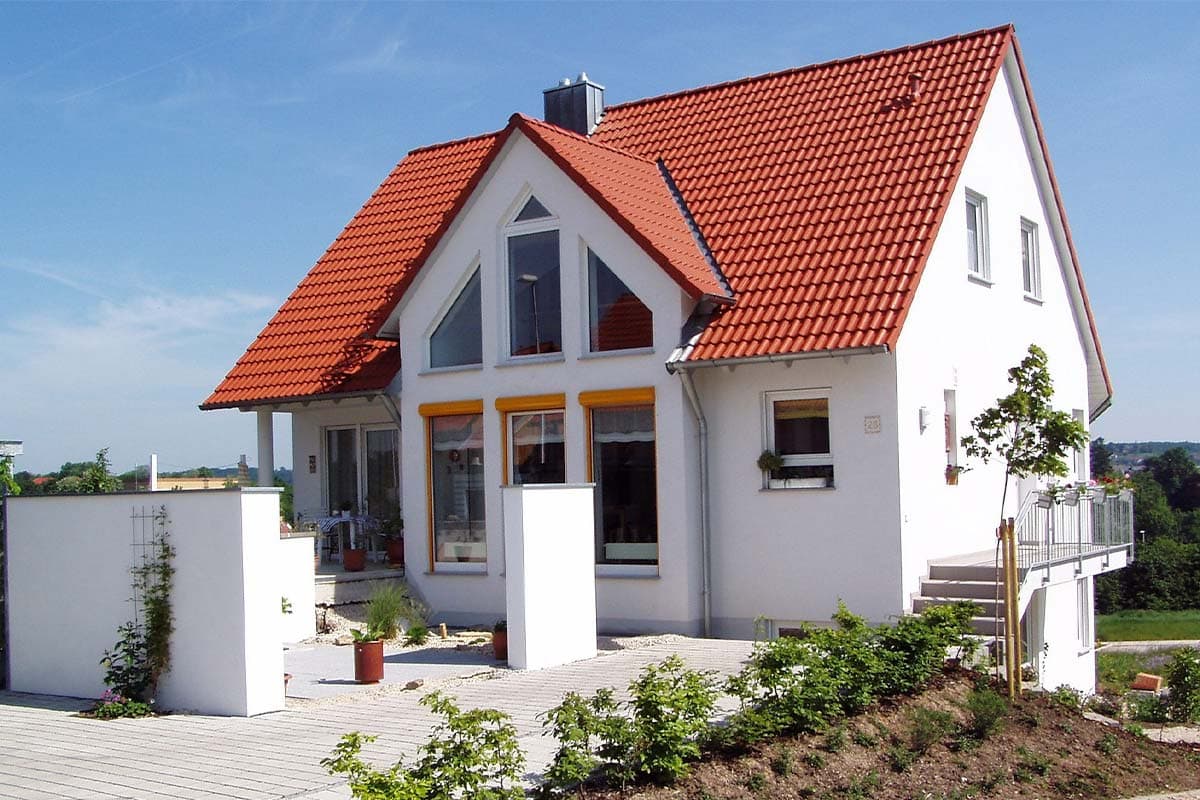 Weißes Einfamilienhaus mit rotem Satteldach, Garten und Terrasse bei sonnigem Wetter.