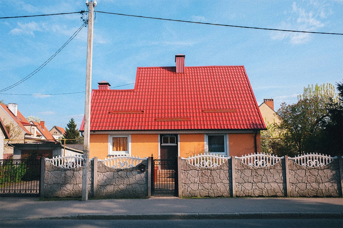 Einfamilienhaus mit rotem Ziegeldach und gelber Fassade hinter einem Steinzaun mit weißen Gittern.