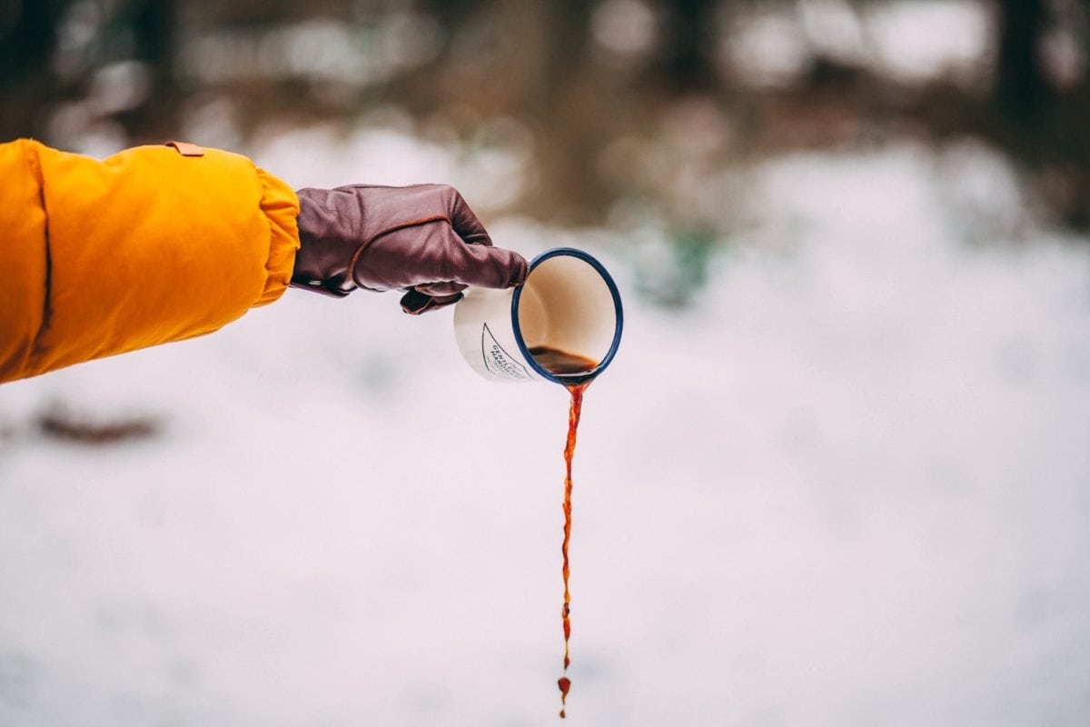 Person gießt Kaffee aus einem Emaille-Becher in den Schnee.