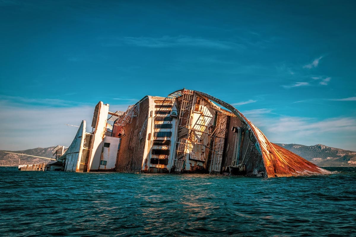 Ein großes, teils untergegangenes Schiff mit rostigen Strukturen ragt aus dem Meer, mit einer Küstenlandschaft im Hintergrund.