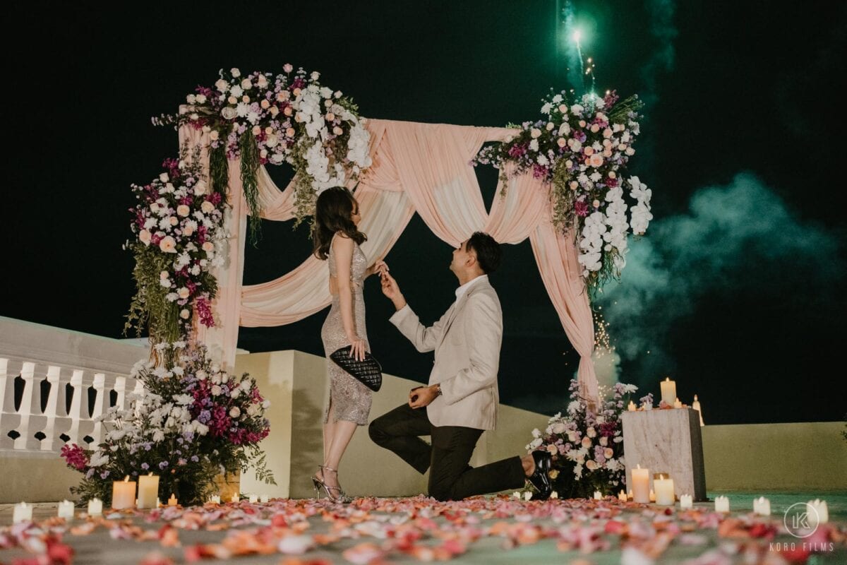 Day kneels to propose to Oil under a flower arch on a riverside terrace at night in Bangkok