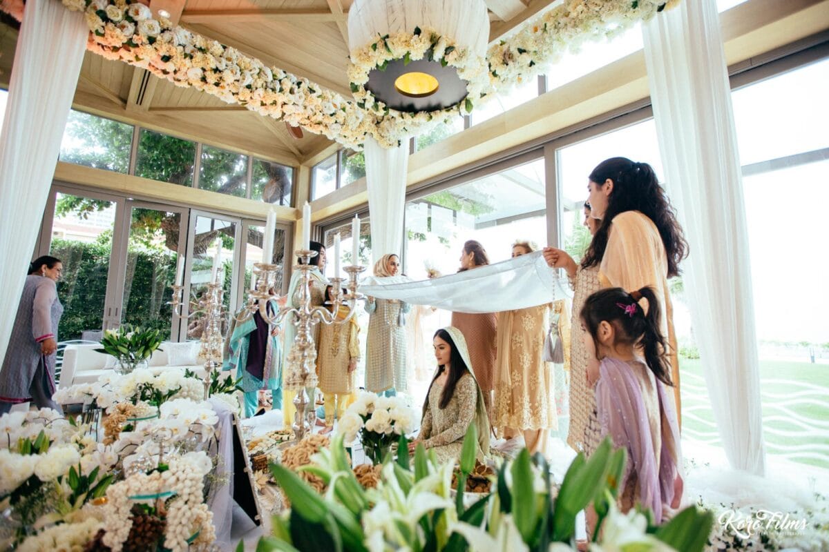 Women of the family hold a white veil above the bride at her Nikah in a glass pavilion in Hua Hin, Thailand
