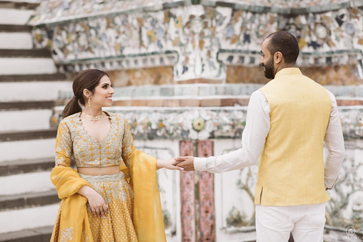 Bride in a mustard lehenga and groom in a yellow Nehru jacket hold hands in front of the porcelain mosaics of Wat Arun, Bangkok