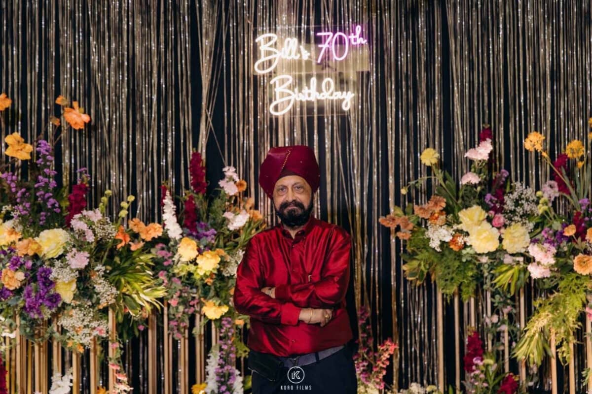 Bill in a red turban in front of a 'Bill's 70th Birthday' neon sign and flower wall at his party in Bangkok