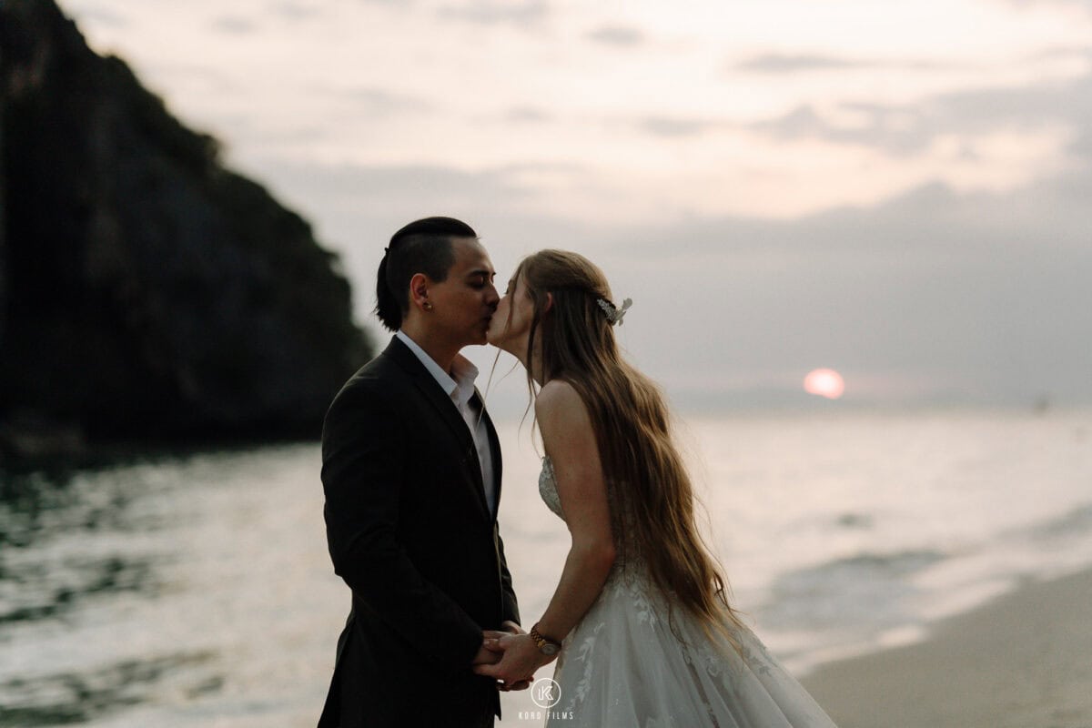 Alessandra and Bruno kiss on the beach at sunset beside the limestone cliffs of Rayavadee, Krabi