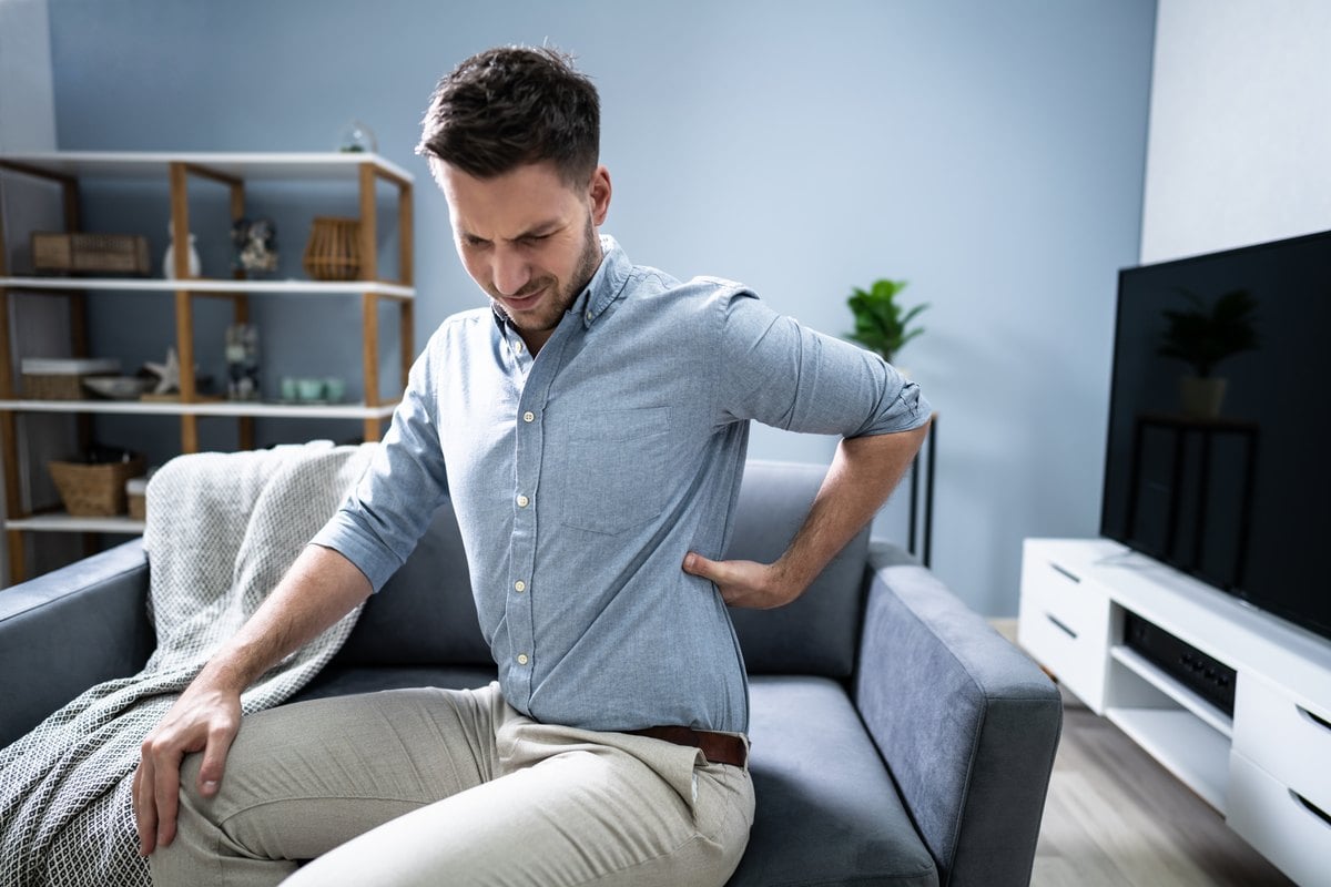 Man sitting on a gray sofa at home, holding his lower back and grimacing in pain