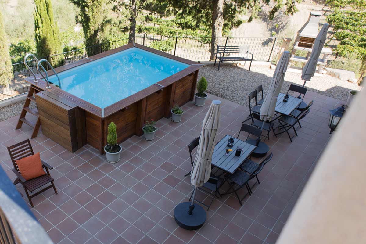 Terrassa on hi ha una barbacoa al fons, una piscina, unes gandules de fusta i taules i cadires amb para-sols, i una escala que puja a mà dreta.