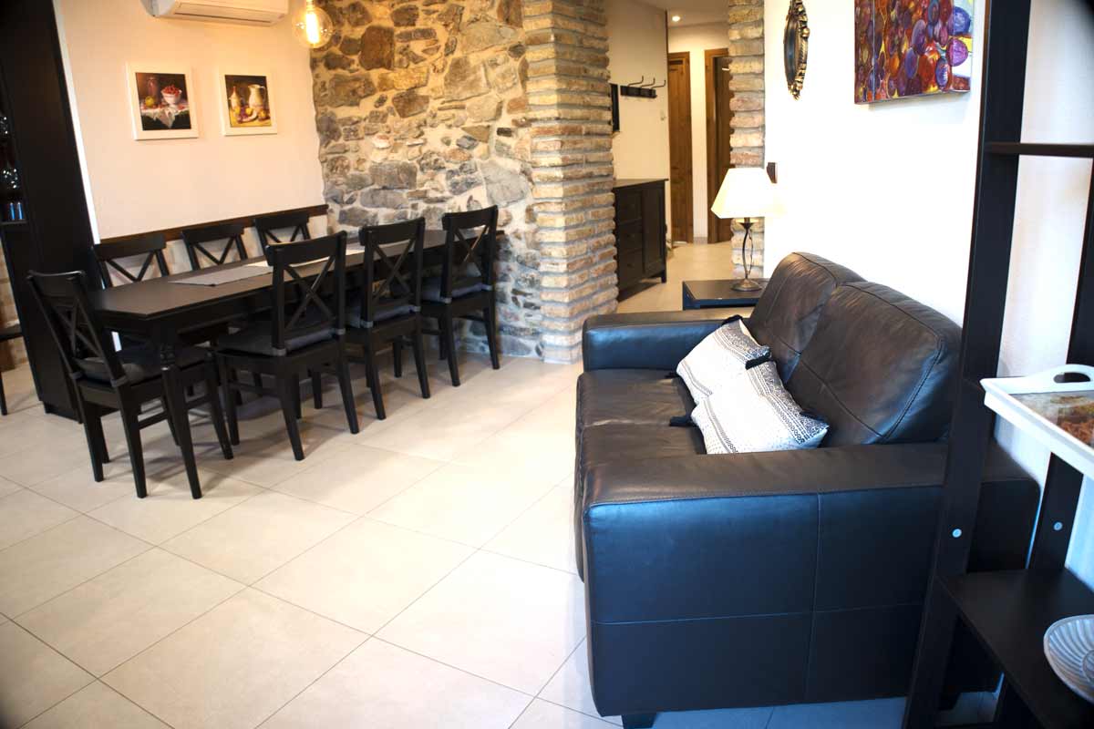 Sala on hi ha una taula llarga i cadires negres, un sofà de pell negra, amb coixins blancs i detalls negres, parets llises i una amb pedra vista.