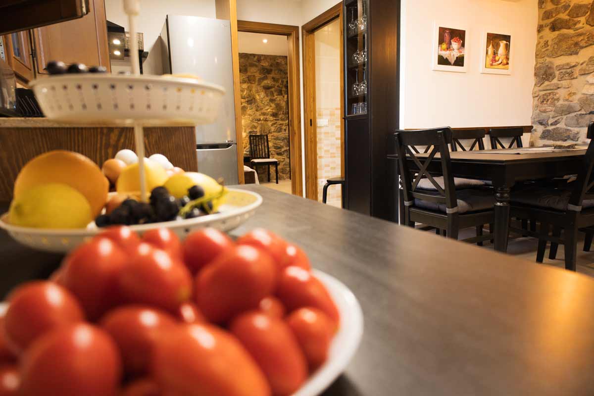 Fotografía detalle donde se ven frutas y verduras desenfocadas y se ve el comedor con la mesa y las sillas de color negro, una pared blanca con cuadros de bodegones hechos a mano colgados y una pared con piedra vista.