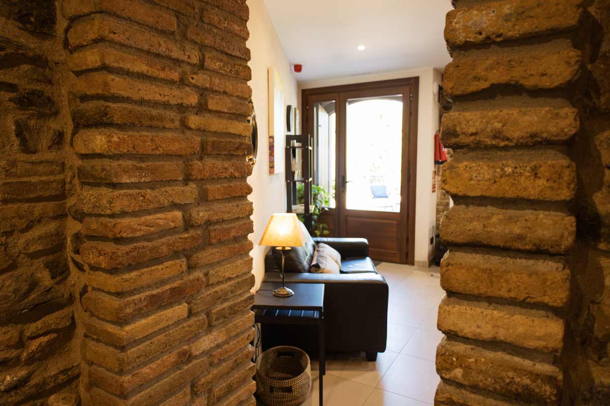 Entrada donde se ve una puerta de madera y un vidrio que hace que entre la claridad de la calle, un sofá negro, una tableta con una luz y un mueble alto negro.