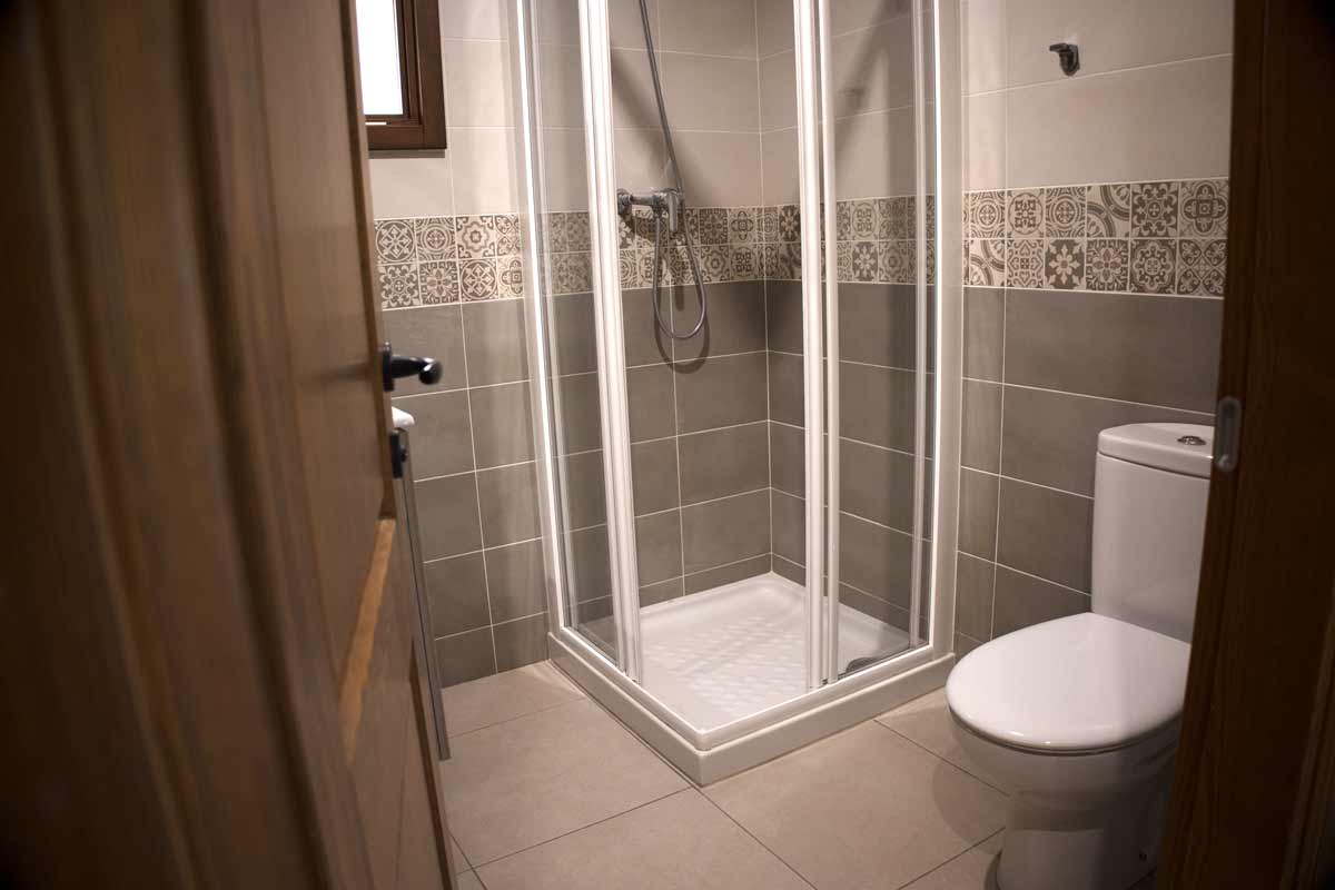 Bany on hi ha un lavabo, una dutxa tancada amb una cristallera vista, una paperera, una pica amb un moble de color fusta clar amb dos calaixos, estanteries, un espill i una tovallola color beix.