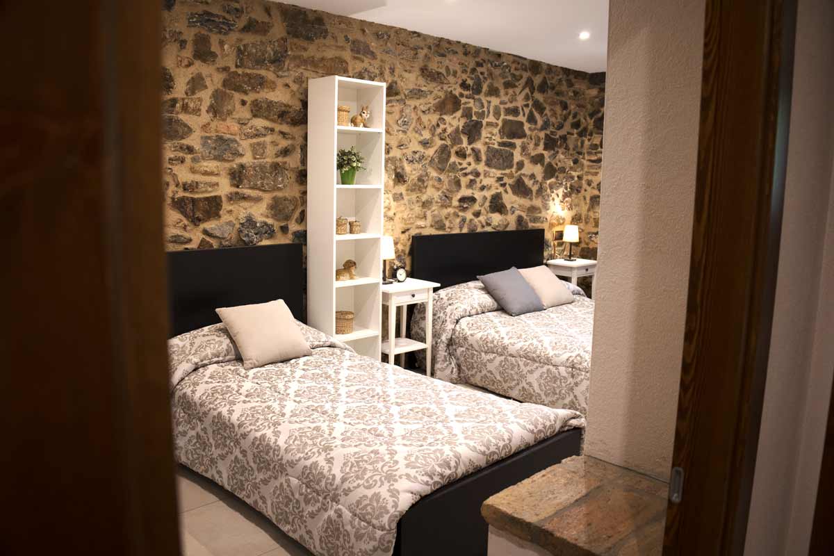 Habitació amb un llit doble i un altre llit individual, amb els mateixos llençols i dos coixins un de color beix i l'altre de color gris. Entre un llit i un altre ho separa un moble alt que té diferents estanteries i dues tauletes de nit.