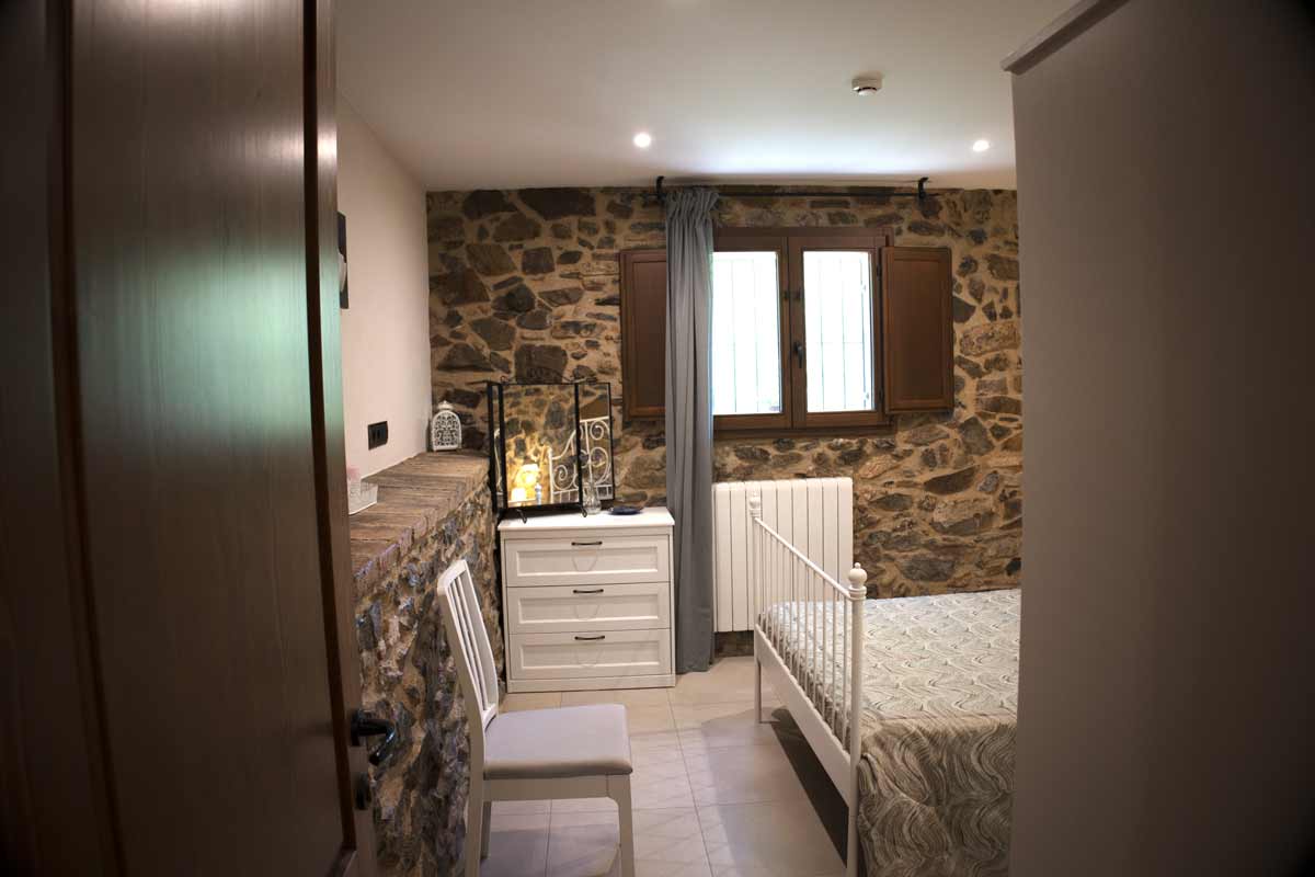 Habitació de matrimoni amb llit doble, un coixí amb una coixinera llisa de color gris, un capçal de ferro pintat de color blanc, un armari blanc amb un espill petit, dues llums a les tauletes de nit i un quadre d'una fotografia on hi ha una flor de color violeta i l'altra de color blanc.