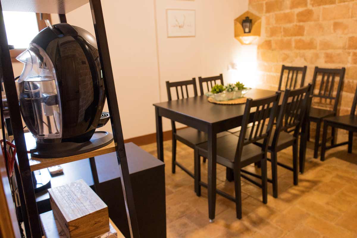 Sala petita amb el terra i les parets de totxanes vistes i una paret llisa pintada de color blanc. Hi ha mobles de color negre, com una taula i cadires, i un armariet amb calaixos i una cafetera de color negre en primer pla.