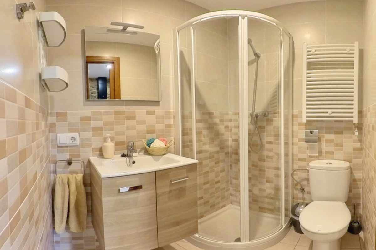 Bany on hi ha un lavabo, una dutxa tancada amb una cristallera vista, una paperera, una pica amb un moble de color fusta clar amb dos calaixos, estanteries, un espill i una tovallola color beix.