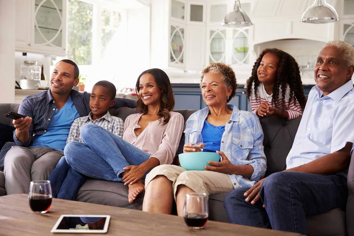 A family of six, spanning three generations, sits on a sofa in a modern living room, relaxed and smiling while watching TV. The room is bright and airy, with a background of windows and white kitchen cabinetry.