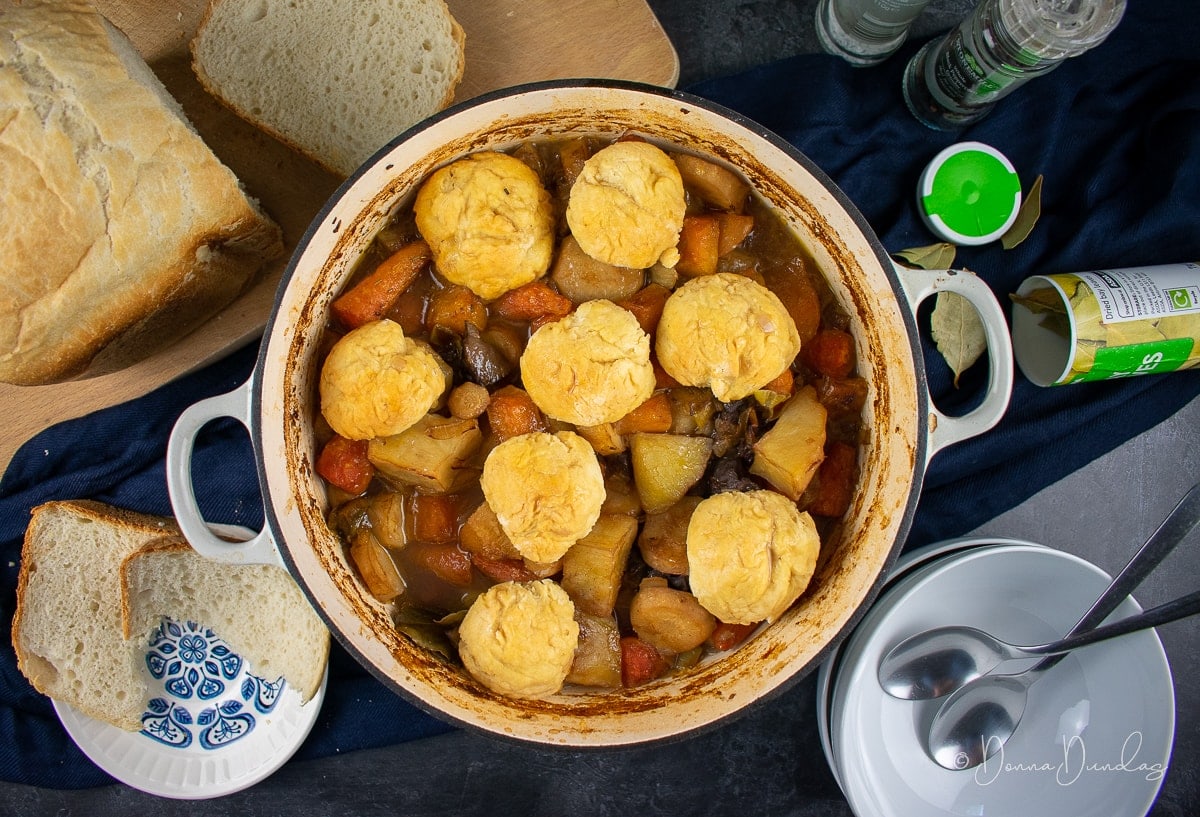 Leftover lamb stew with dumplings in a cast-iron cooking pan. Loaf of bread on chopping board above.