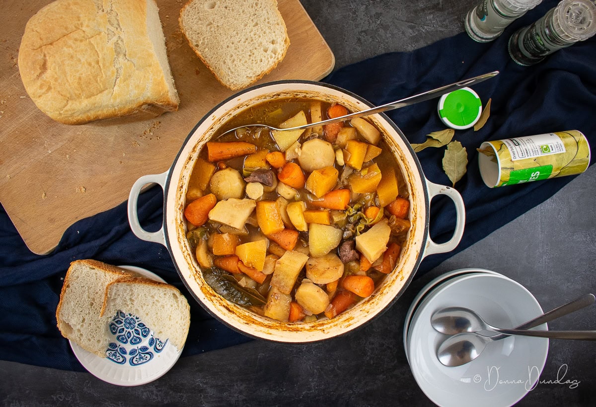 Leftover lamb stew with dumplings in a cast-iron cooking pan. Loaf of bread on chopping board, salt Pepper and bay leaves to the side. Empty bowls at the bottom.