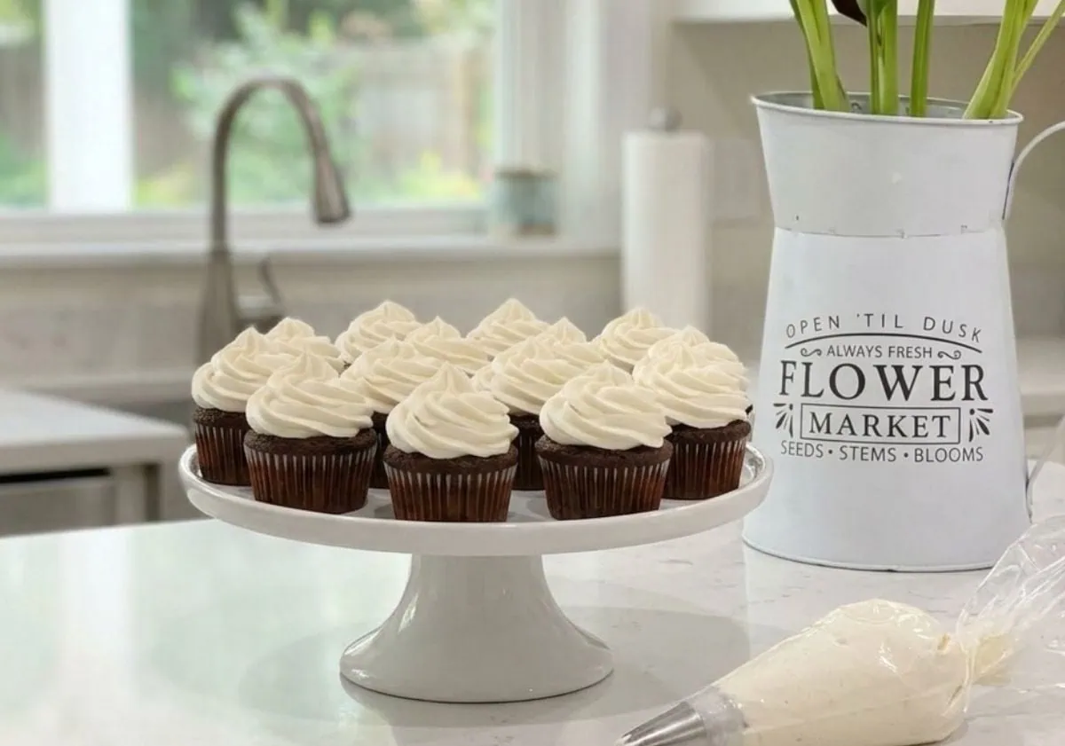 A white cake stand holding several chocolate cupcakes topped with a thick, swirl of fluffy vanilla frosting made without powdered sugar, sitting on a white kitchen counter.