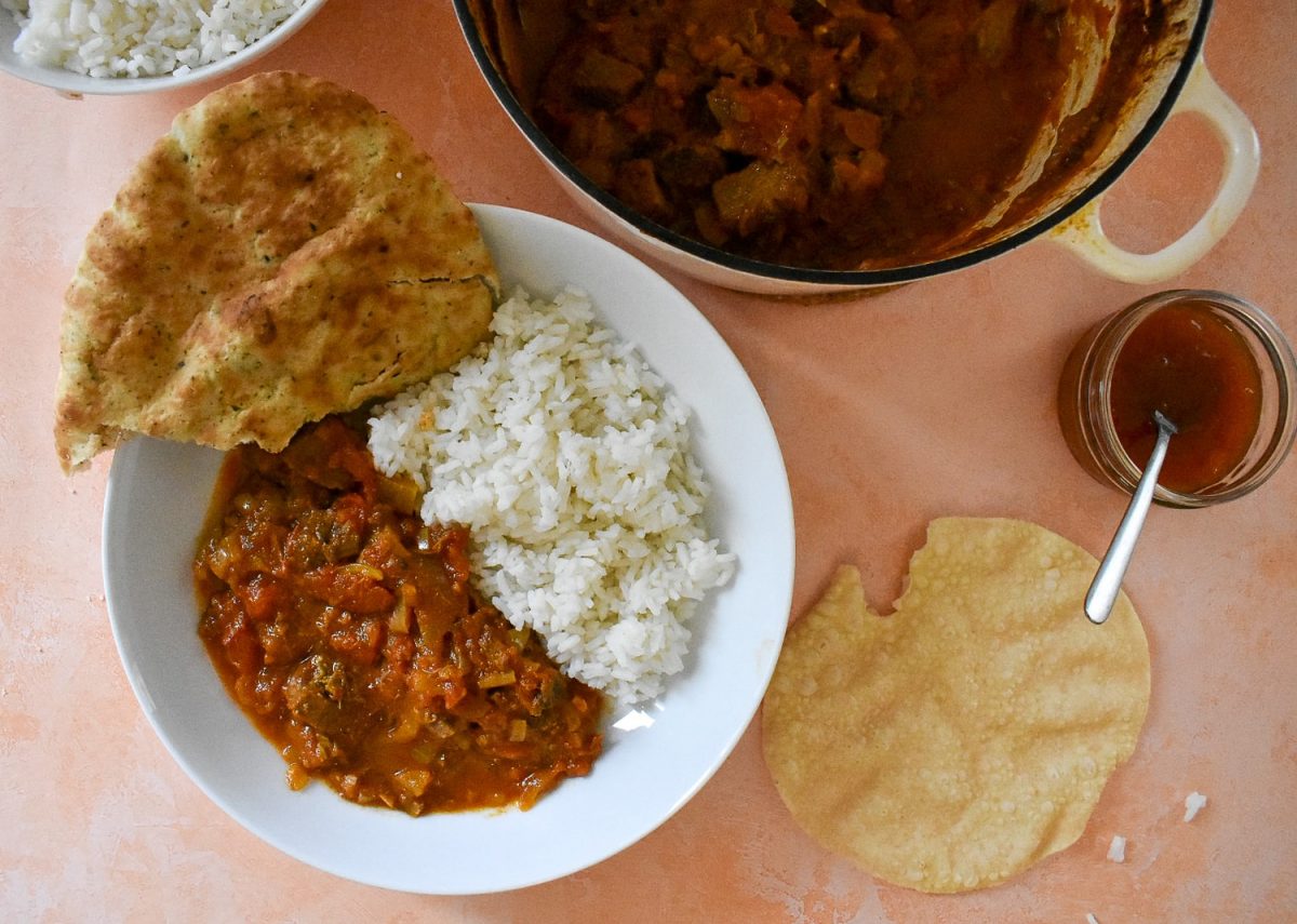 Bowl of served curry with rice. Naan and poppadoms round side, serving bowl leaving shot to top.