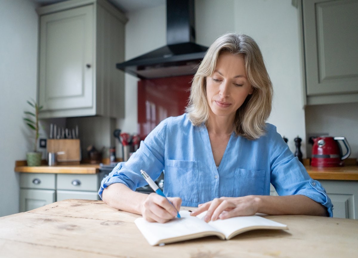 Woman at a wooden kitchen table writing in an open journal with a pen